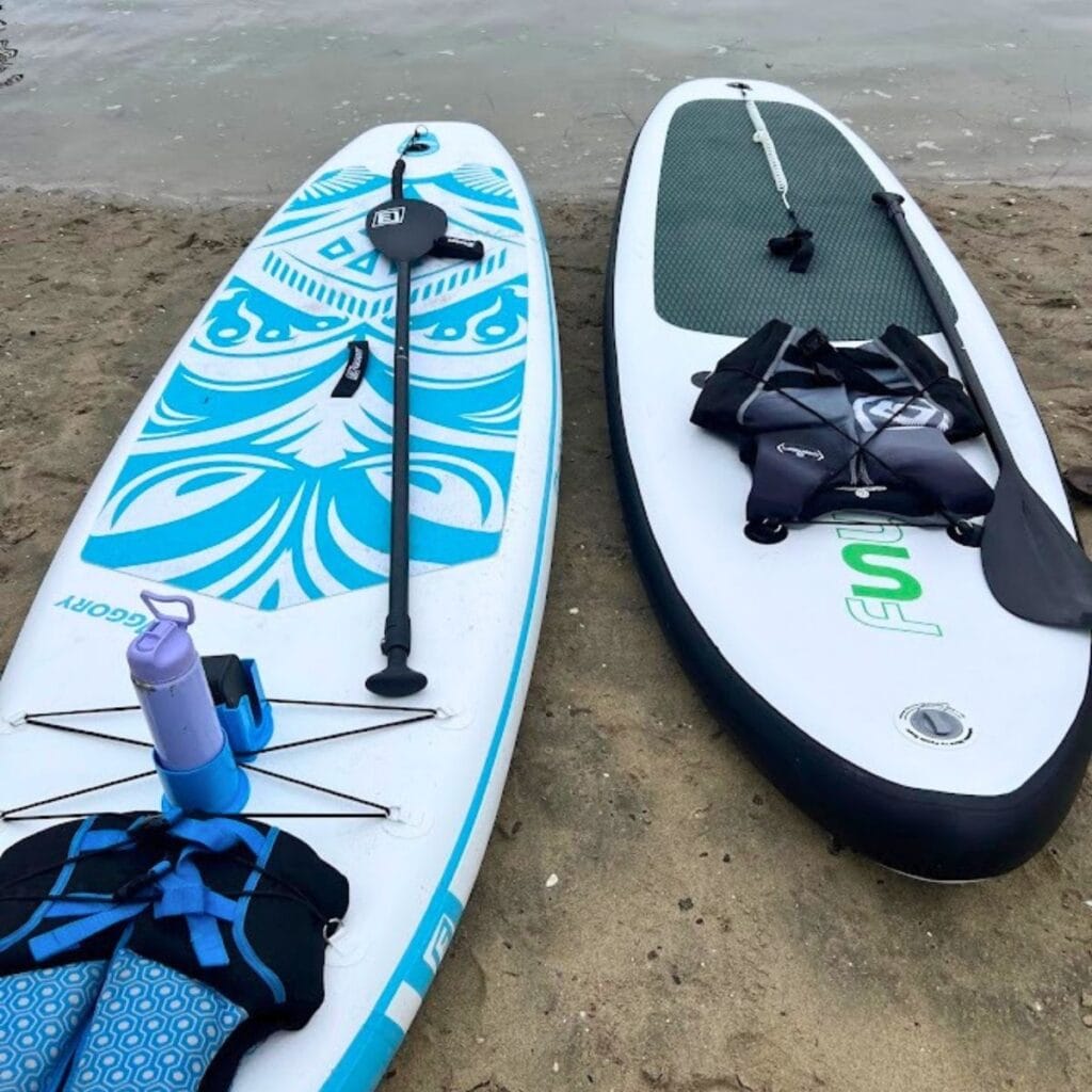 Two stand-up paddleboards on a sandy beach with paddles and gear laid out, ready for a session by calm water.