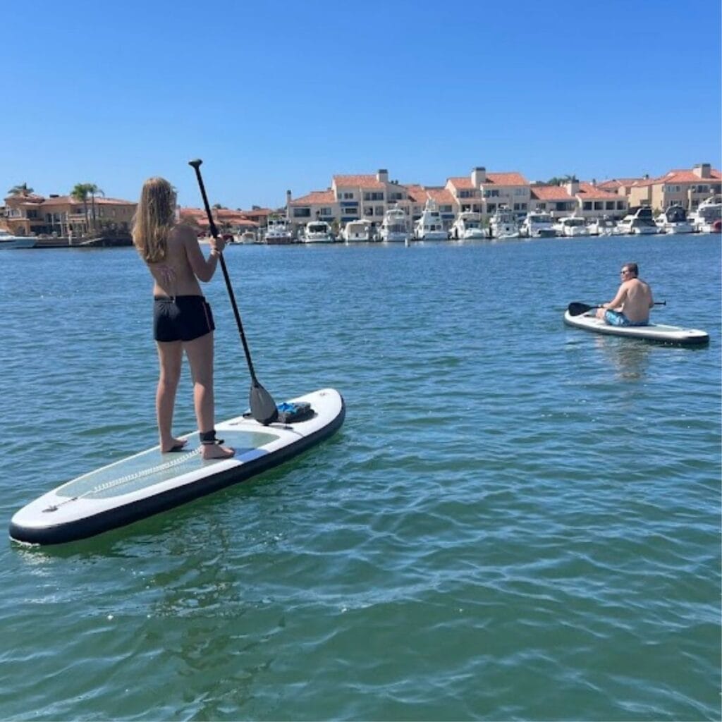 Two people stand-up paddleboarding on calm water near a marina with moored boats and waterfront houses in the background.